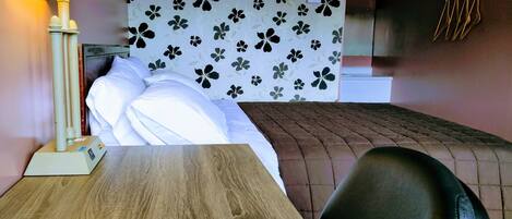 A hotel room with a bed, a wooden table, a lamp, and a wall with a floral pattern.