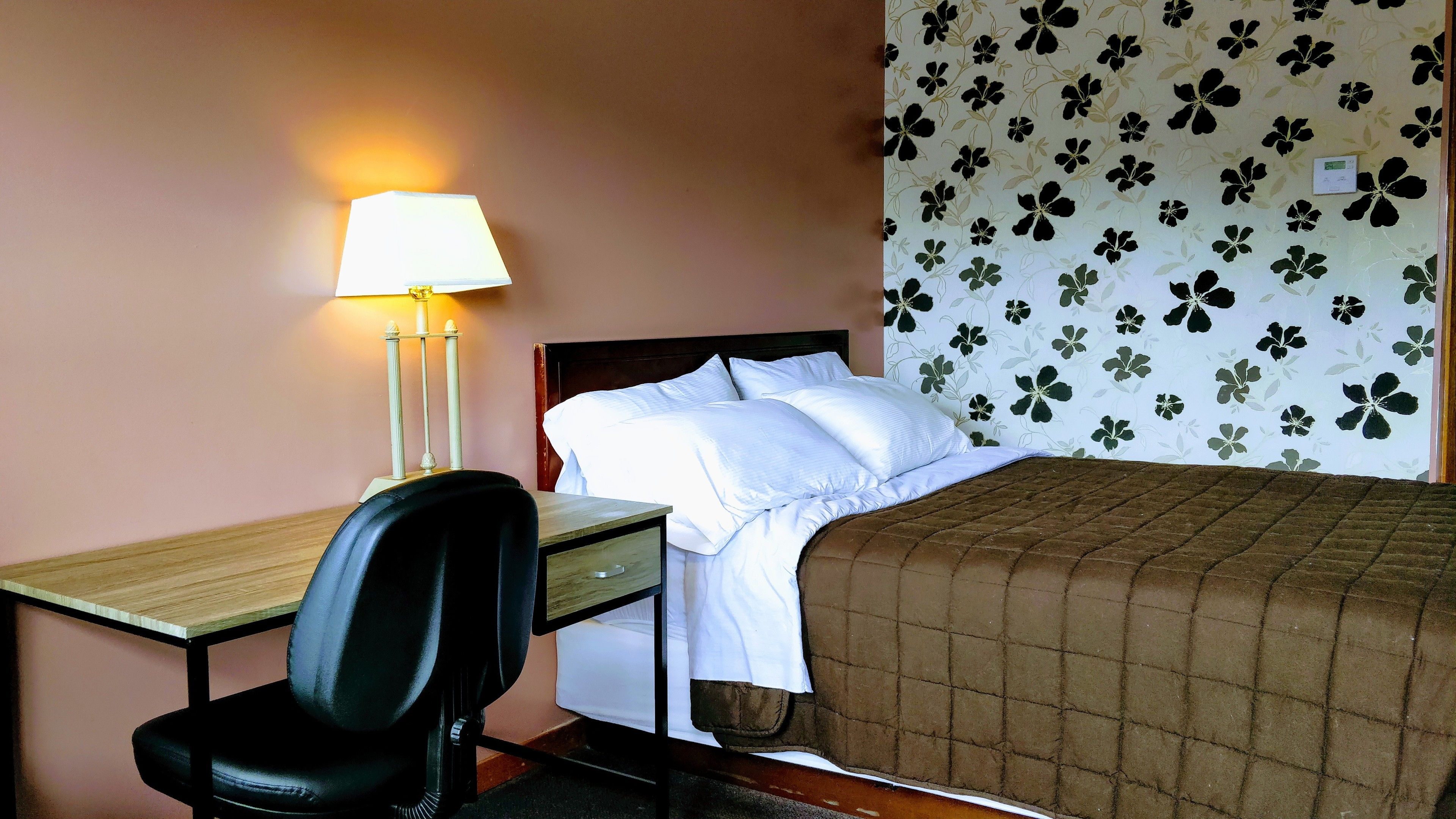 Una habitación de hotel con una cama, un escritorio con una silla, una lámpara y un papel tapiz floral.