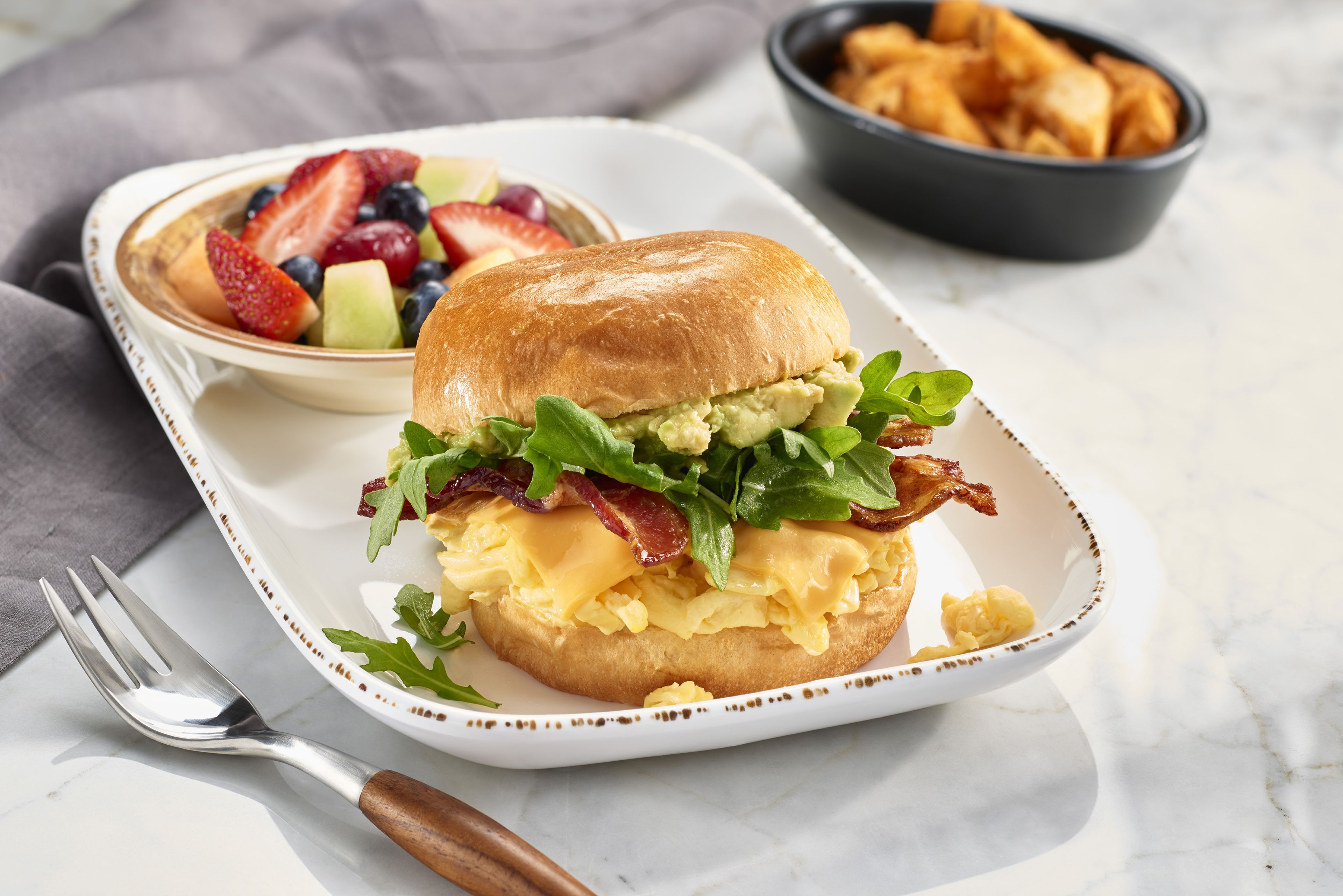 Un plato de desayuno con un sándwich, un tazón de fruta y un tazón de papas fritas.