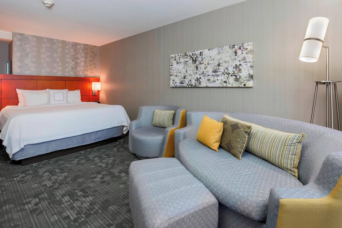 A hotel room with a bed, a grey sofa, and a small armchair. There is a painting on the wall and a floor lamp.