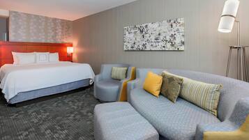 A hotel room with a bed, a grey sofa, and a small armchair. There is a painting on the wall and a floor lamp.