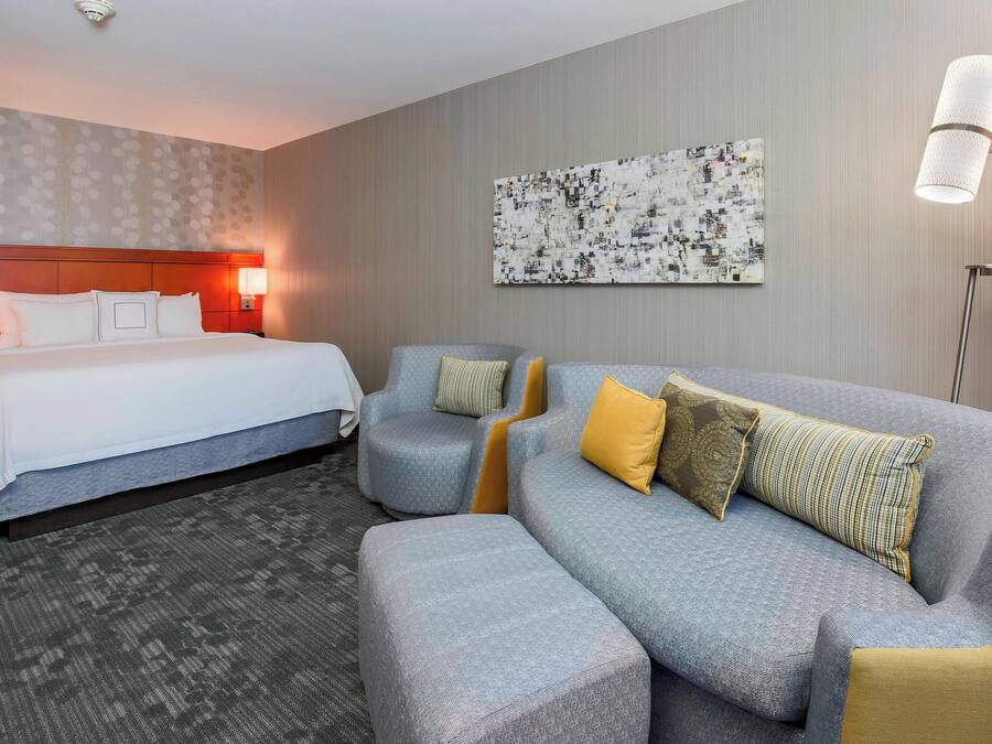 A hotel room with a bed, a grey sofa, and a small armchair. There is a painting on the wall and a floor lamp.
