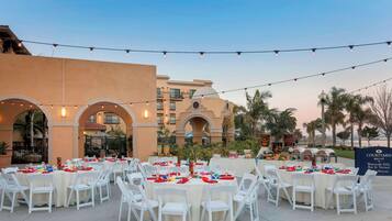 Un área de comedor al aire libre con mesas redondas y sillas blancas, luces colgantes y un edificio con arcos al fondo.