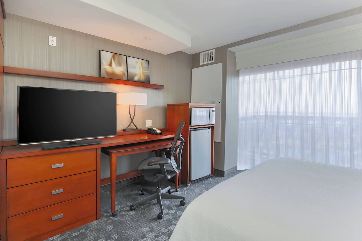 A hotel room with a bed, a desk with a TV, a chair, a mini-fridge, and a large window with sheer curtains.