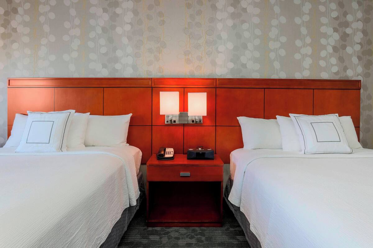 A hotel room with two beds, a wooden headboard, a nightstand with a phone and radio, and a patterned wallpaper.