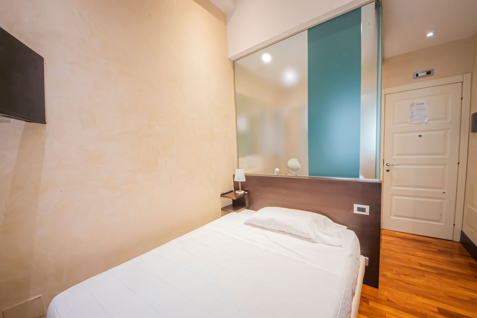 A hotel room with a bed, a nightstand, a mirror, a door, and a wooden floor.