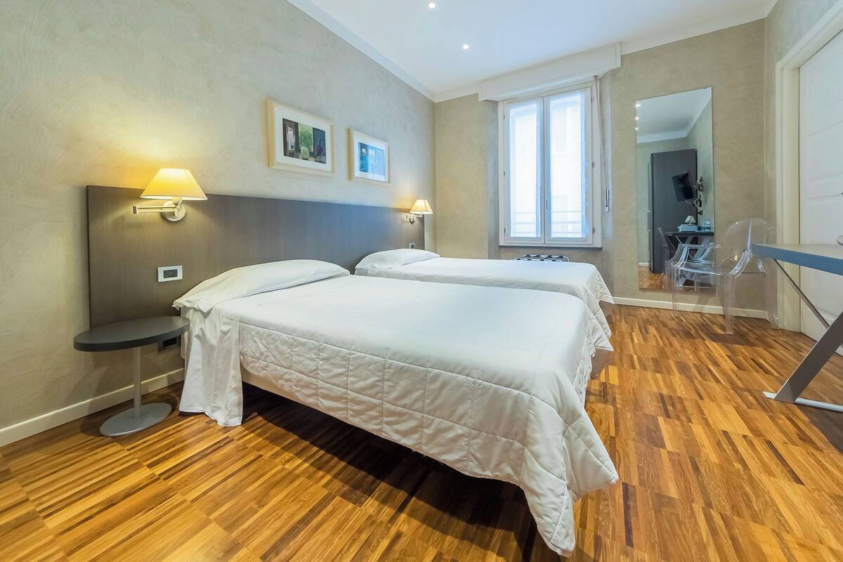 A hotel room with two beds, a desk, a chair, a mirror, and a window.