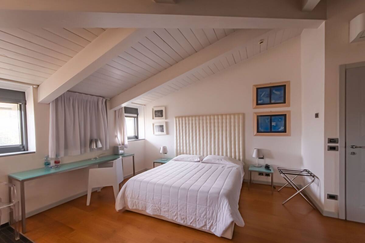 A bedroom with a large bed, a desk, and a chair. There are two windows with curtains, a door, and a wall-mounted shelf with pictures.