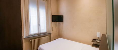 A bedroom with a bed, a radiator, a window with curtains, a TV mounted on the wall, and a bedside table with a lamp.
