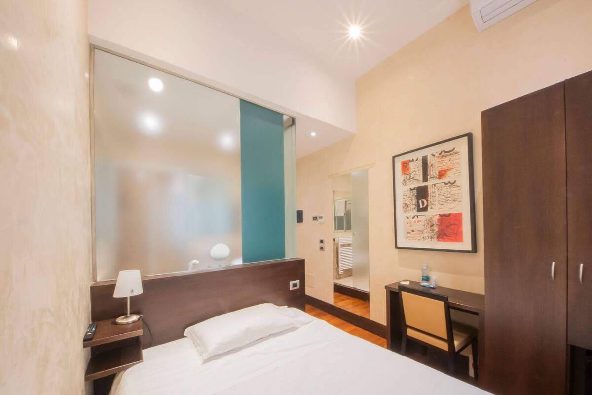 A hotel room with a bed, a desk, a chair, a mirror, and a framed artwork on the wall.