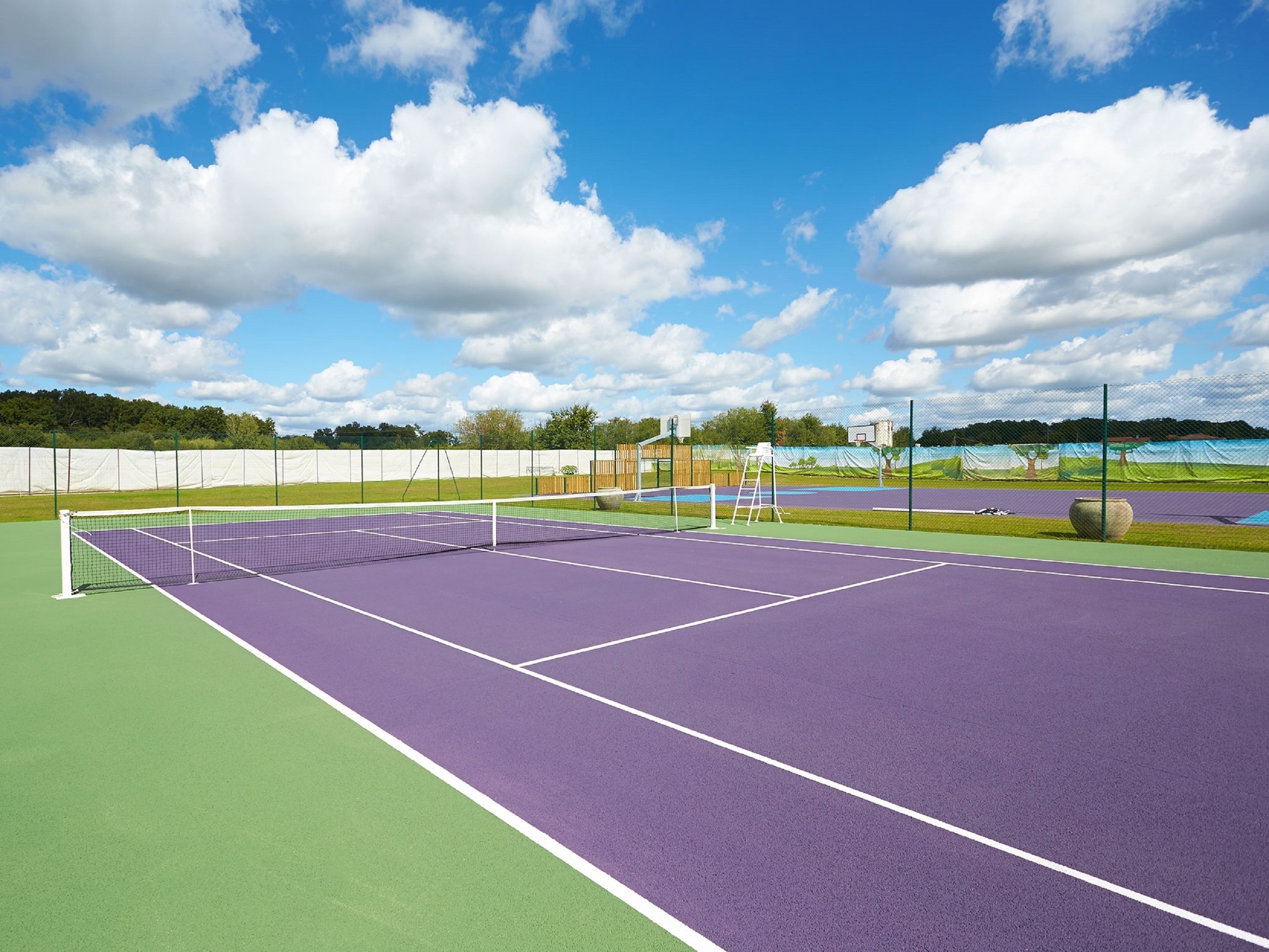 Een tennisbaan met een net, omgeven door een hek, en een basketbalring in de verte.