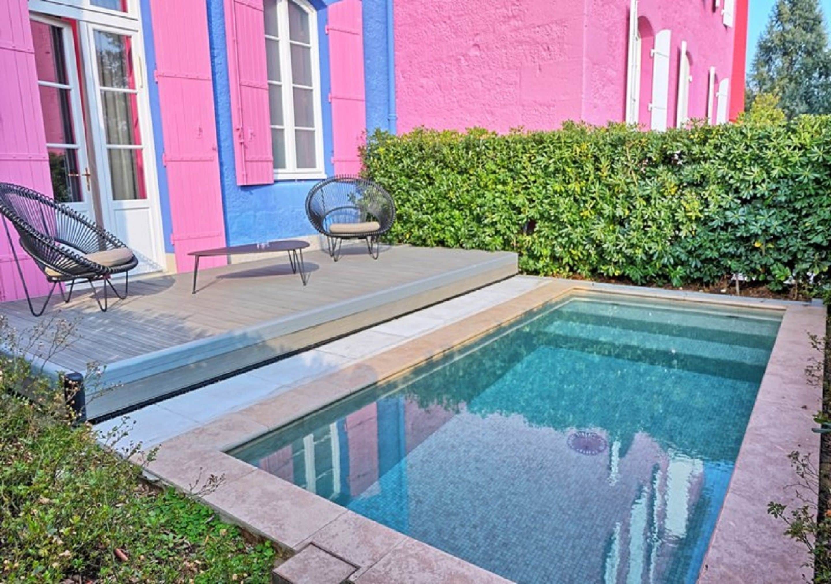 A swimming pool with a wooden deck, two chairs, and a pink and blue building in the background.