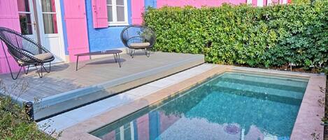A swimming pool with a wooden deck, two chairs, and a pink and blue building in the background.