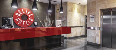A hotel reception area with a red counter, black pendant lights, a digital display, and a sign reading "BELIEVE HOTEL".