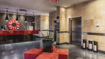Zona de recepción de un hotel con un mostrador rojo, un reloj digital y un ascensor.