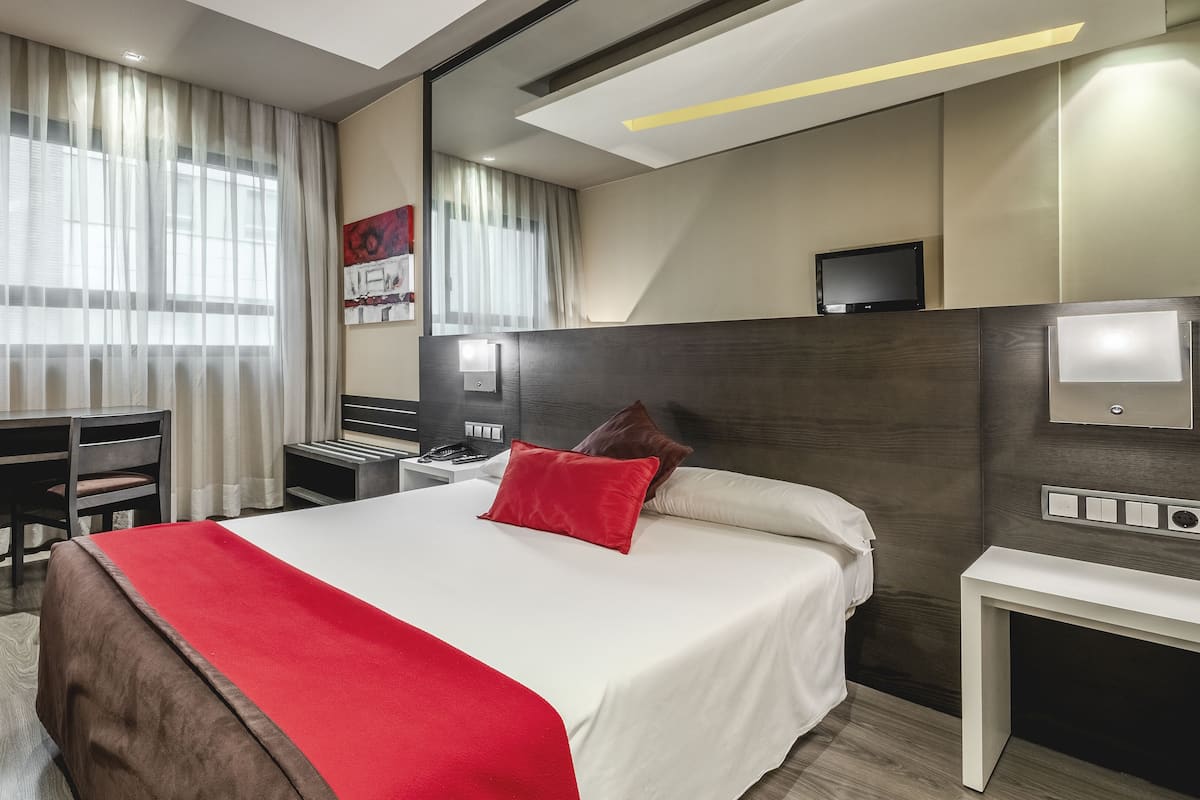 A modern hotel room with a large bed, a desk, and a chair. There is a TV on the wall and a window with curtains.