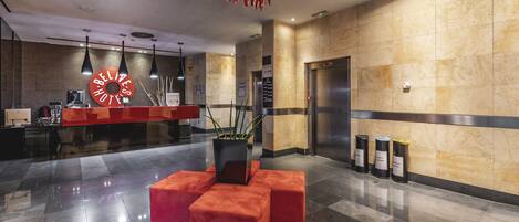 A modern lobby with a reception desk, a red clock, a potted plant, and a trash bin.