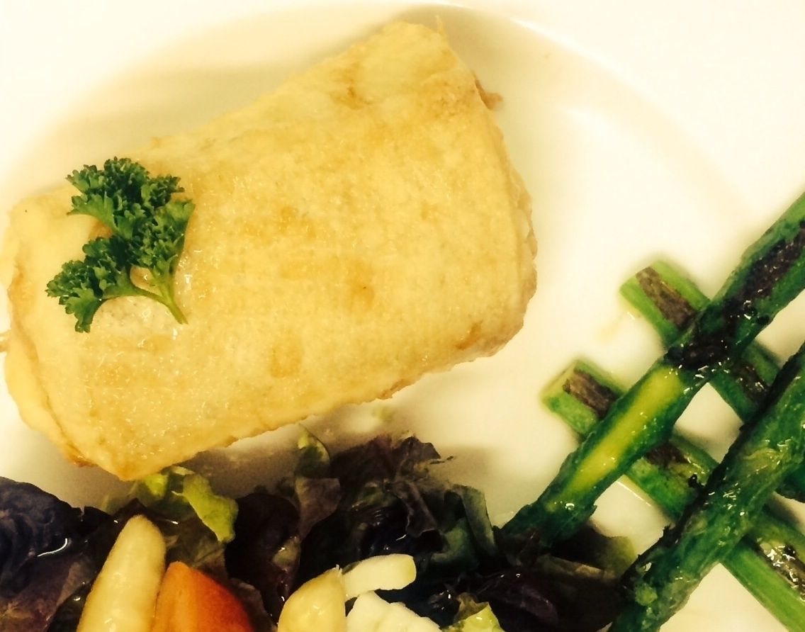 Um prato com um alimento empanado, possivelmente peixe, decorado com salsa, acompanhado de legumes variados, incluindo aspargos e verduras.