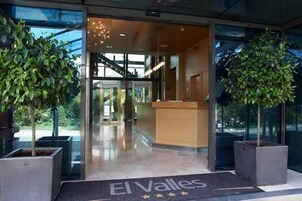 Entrada de un hotel con mostrador de recepción, puertas de cristal y plantas en macetas.