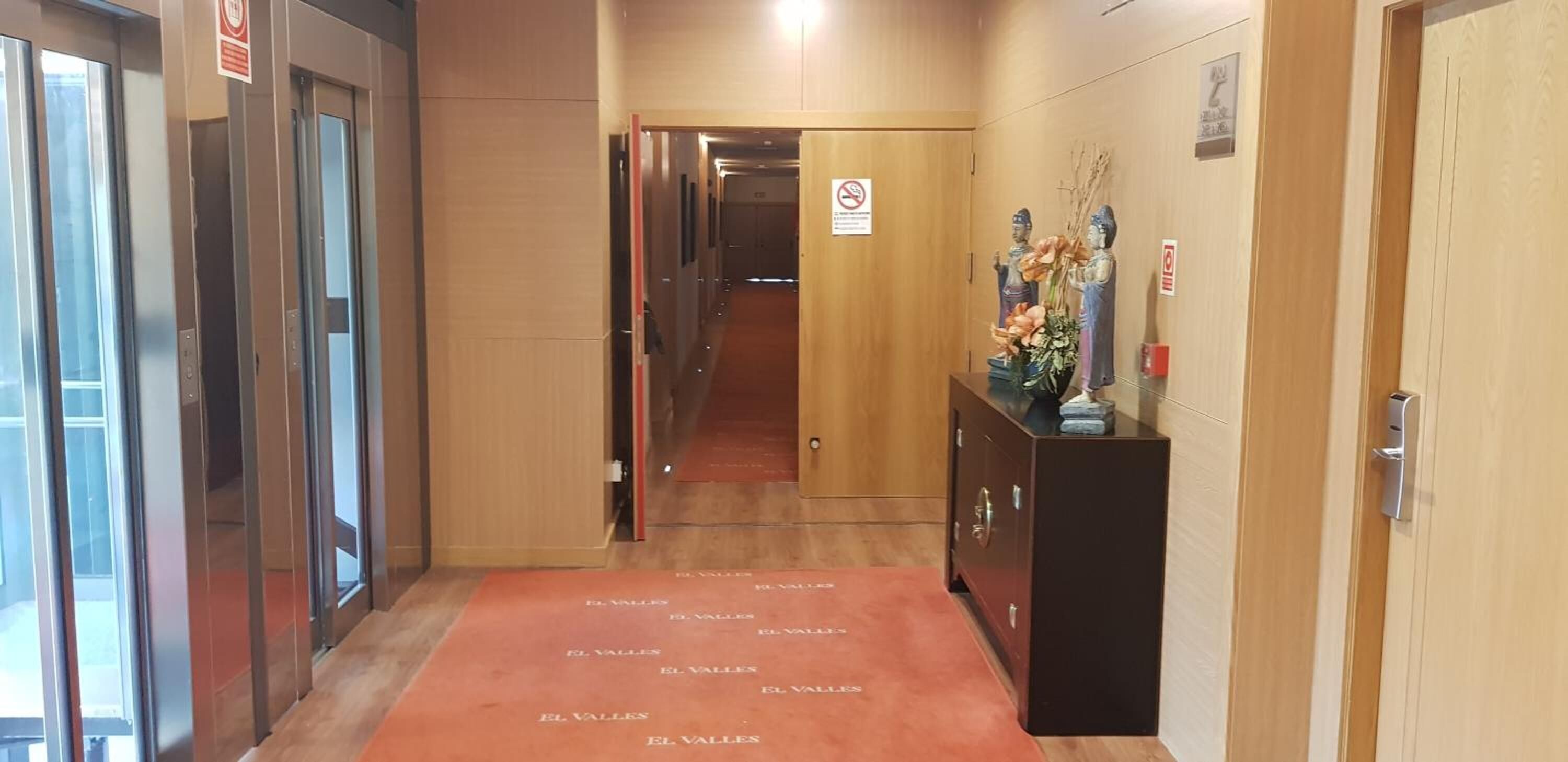 Corridoio di un hotel con ascensori, una porta di legno e una reception con un vaso di fiori.