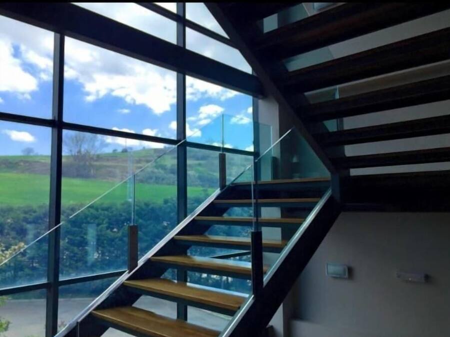 Una escalera moderna con barandillas de vidrio que conduce a un balcón con vistas a un paisaje verde.