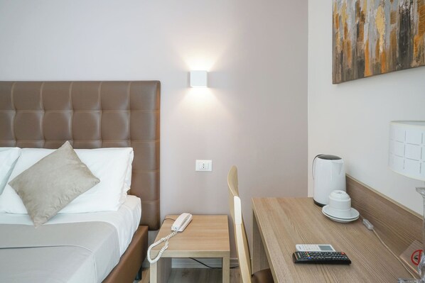 Quarto de hotel com cama, mesa de cabeceira, telefone, cafeteira e um quadro na parede.