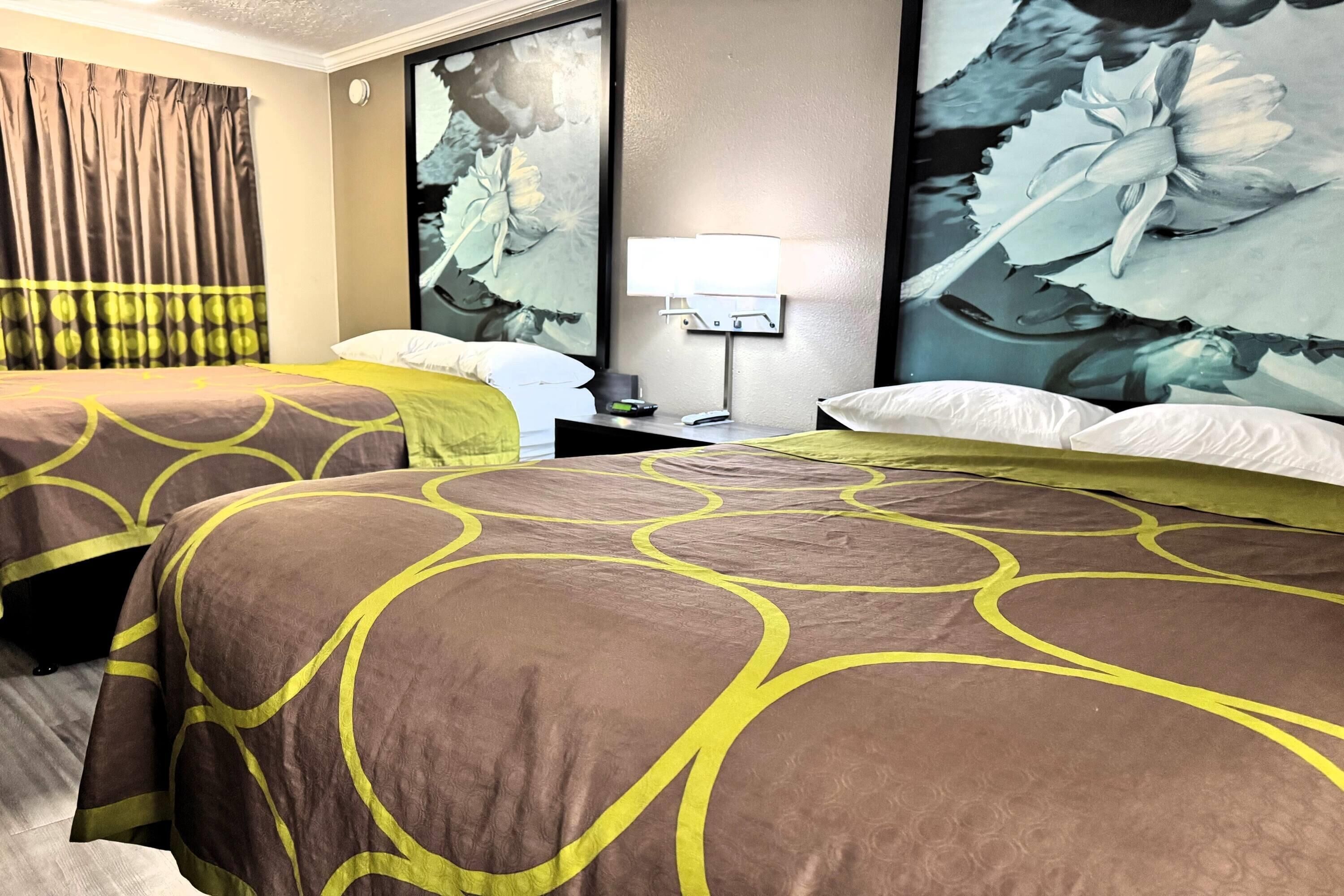 A hotel room with two beds, a nightstand, a lamp, and a large floral artwork on the wall.