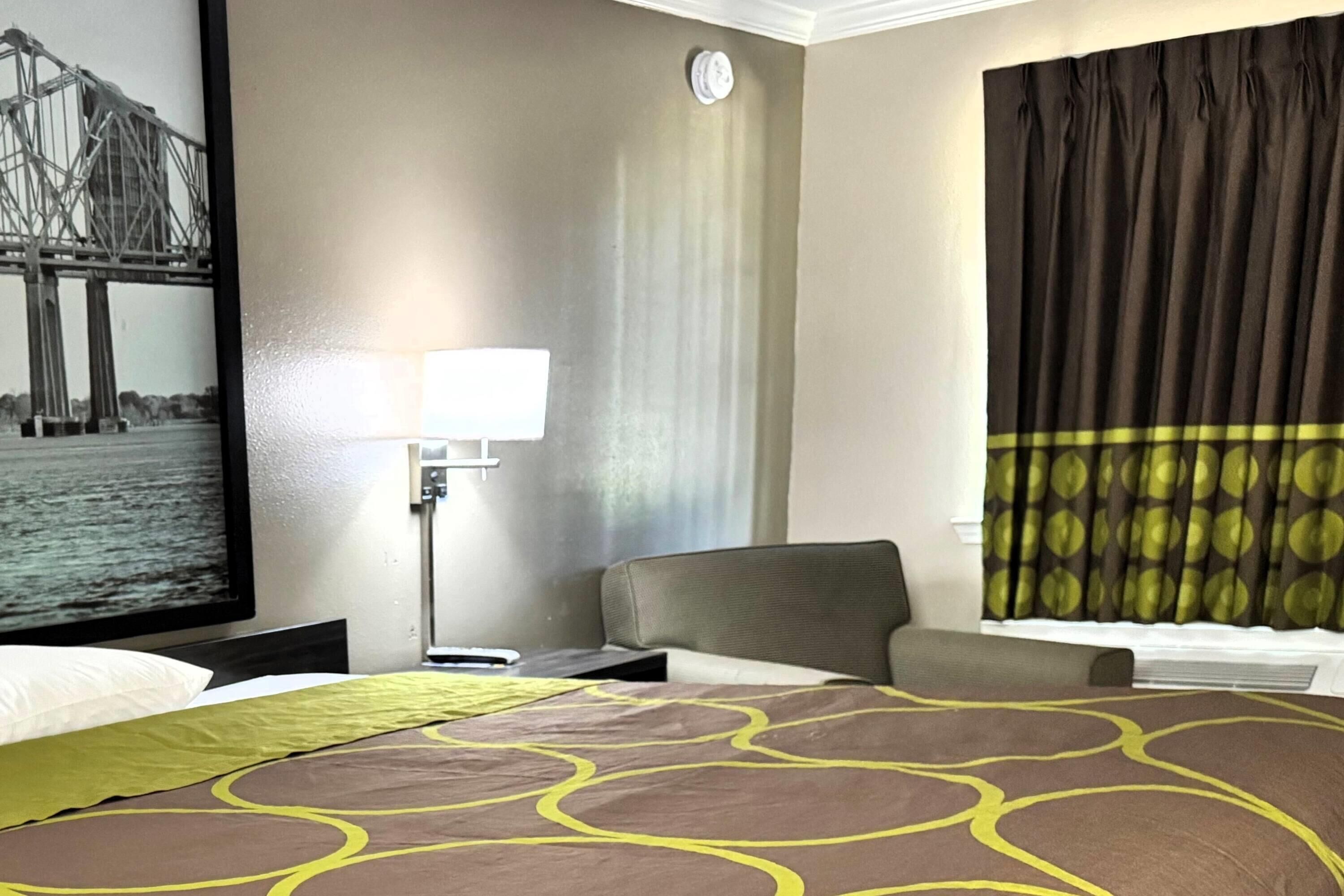 Une chambre d’hôtel comprenant un lit, une table de chevet, une lampe, une grande fenêtre avec des rideaux et une horloge fixée au mur.