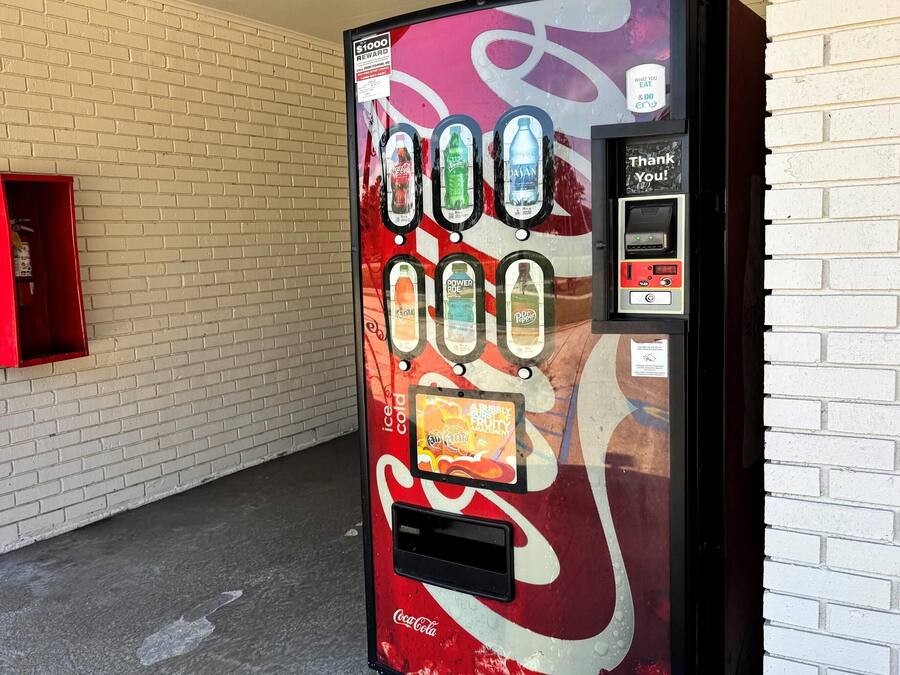 A Coca-Cola branded vending machine with a reward program sign, located next to a white brick wall.