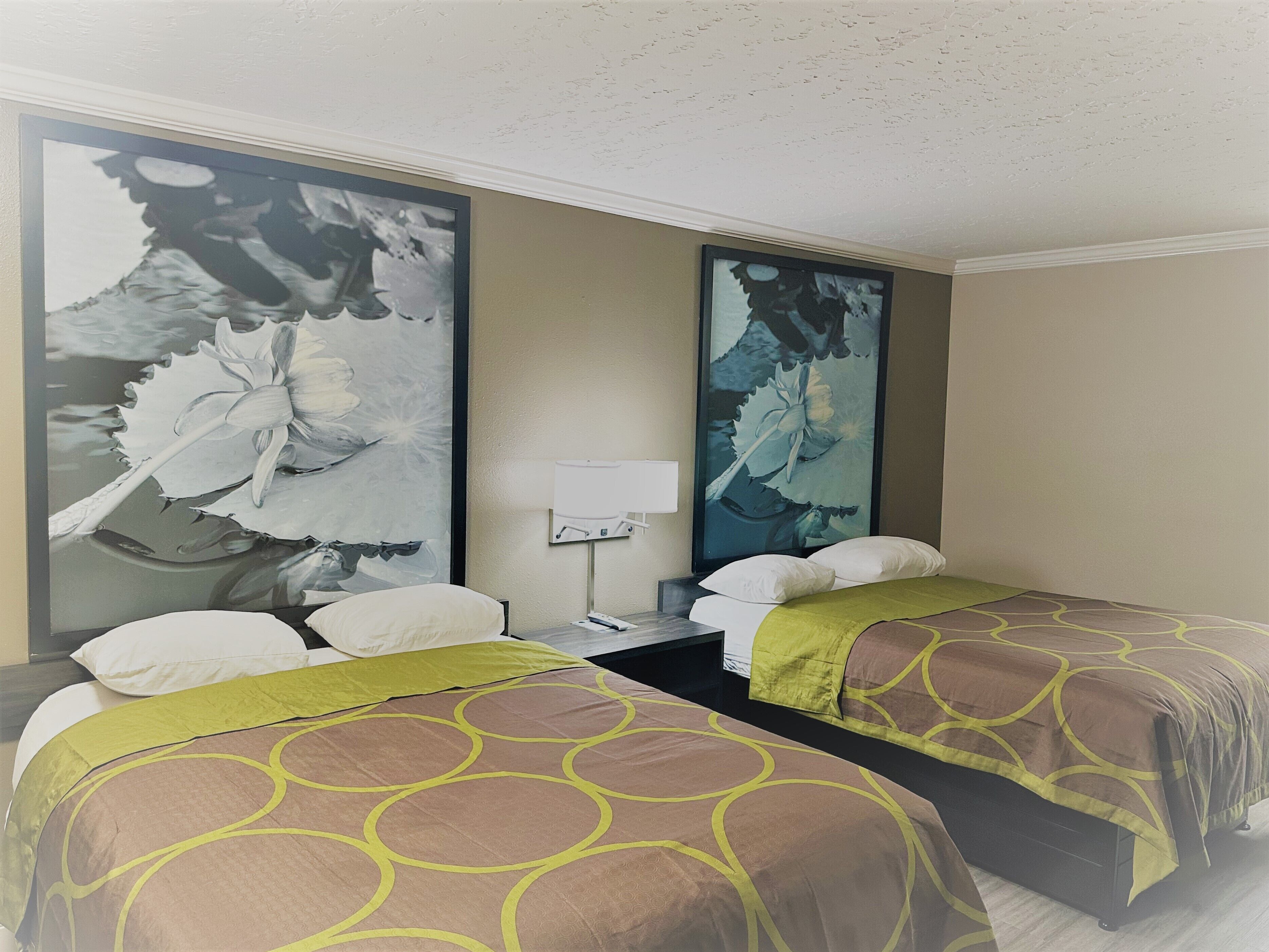 A hotel room with two beds, each with a brown and yellow patterned bedspread, a nightstand with a lamp, and large floral artwork on the walls.