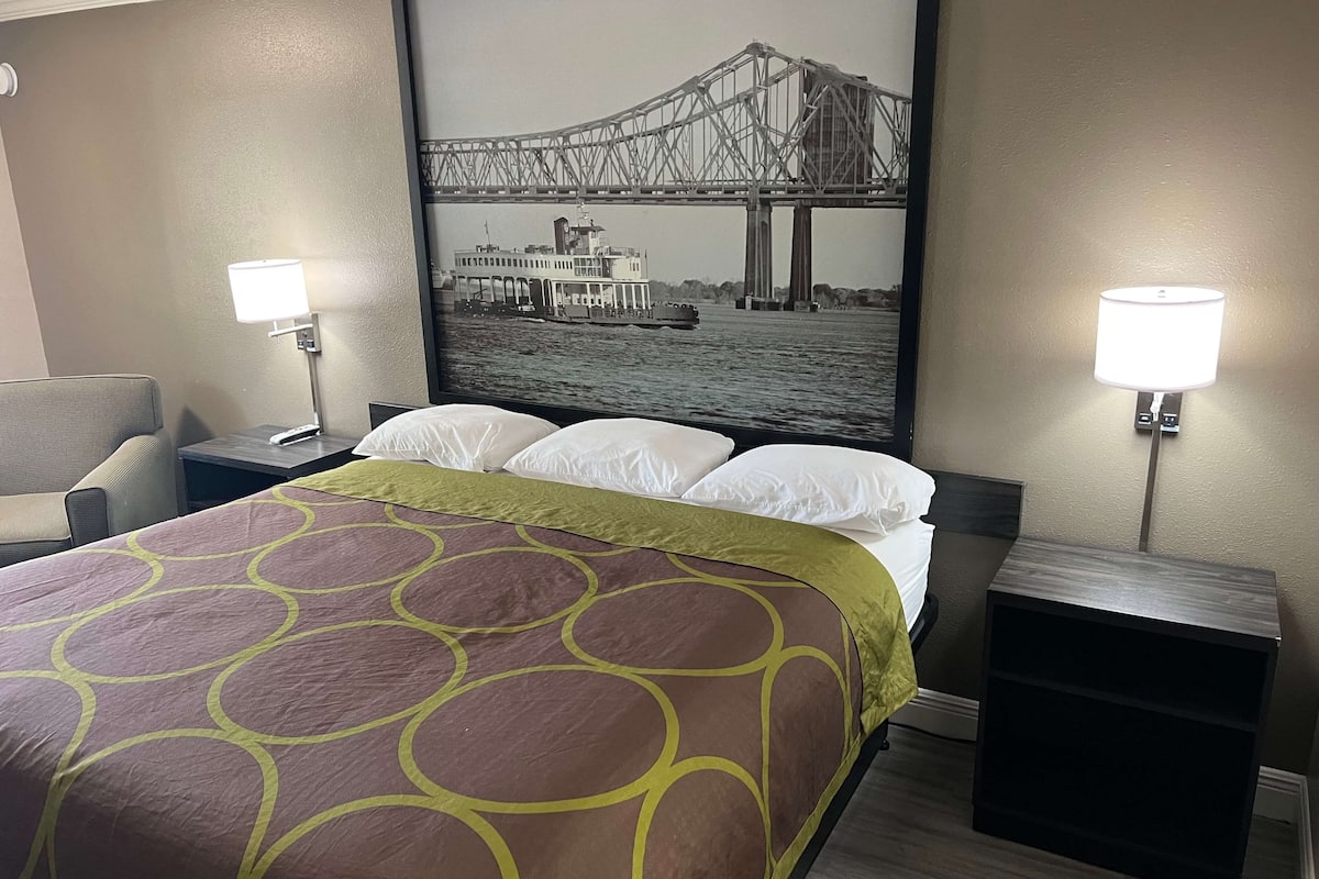 Ein Hotelzimmer mit einem Bett, zwei Nachttischlampen, einem Kopfteil, einem Nachttisch und einem großen gerahmten Bild von einer Brücke und einem Fluss.