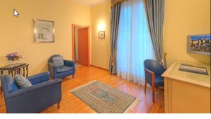 Un salón con dos sillones azules, una mesita con un jarrón, una alfombra y un escritorio con un marco de fotos.