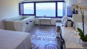 A hotel room with a large window, a bathtub, a small table with a vase of flowers, and a bed with a white bedspread. - Astarte Suites (Santorini)
