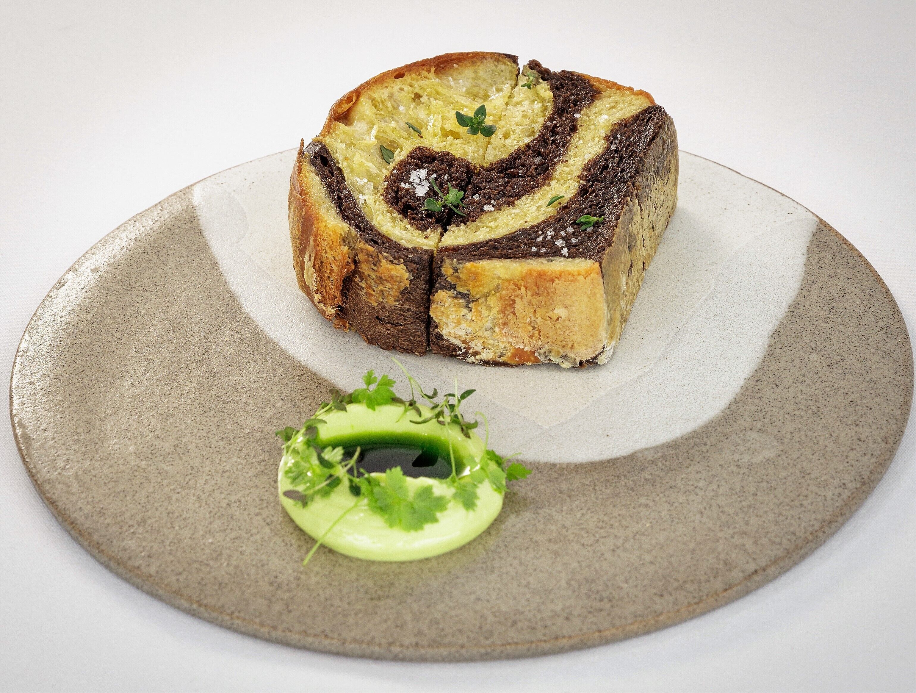 Une tranche de pain décorée d’un motif en spirale, présentée sur une assiette et accompagnée d’une petite garniture verte.