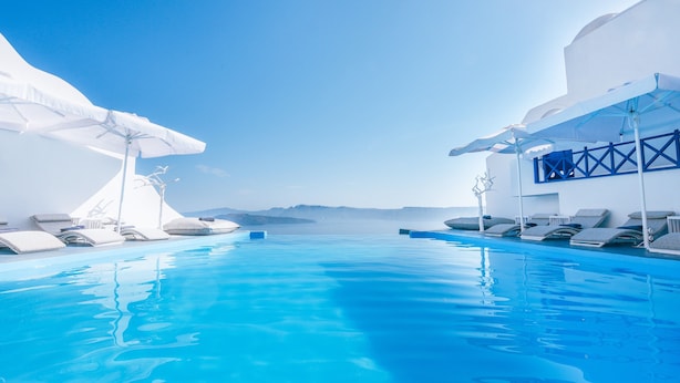 A clear blue infinity pool with white lounge chairs and umbrellas, surrounded by white buildings with a view of the sea and distant mountains. - Astarte Suites (Santorini)