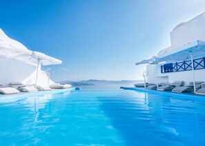 A clear blue infinity pool with white lounge chairs and umbrellas, surrounded by white buildings with a view of the sea and distant mountains. - Astarte Suites (Santorini)