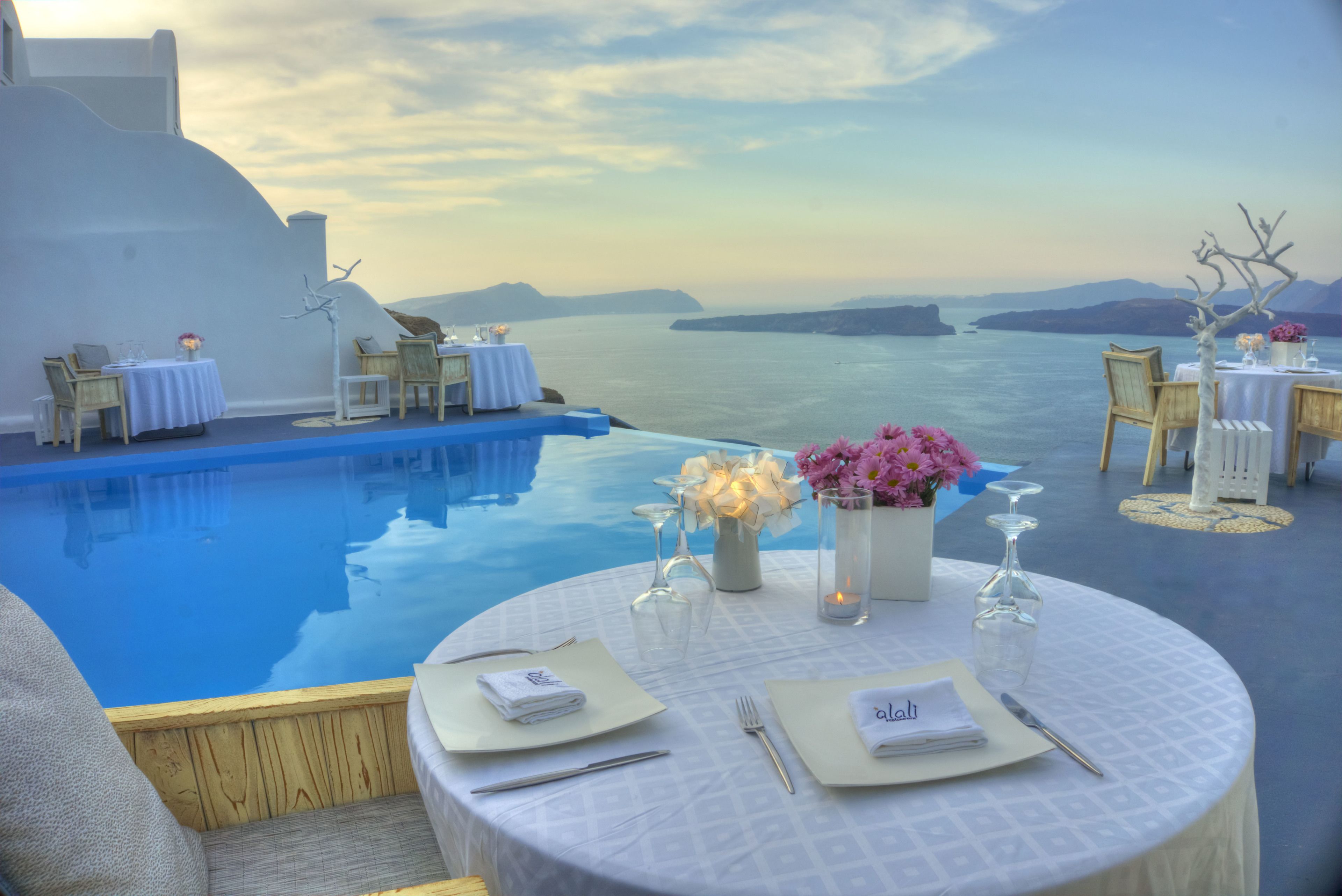A dining area with round tables set for two, overlooking a pool and the sea.