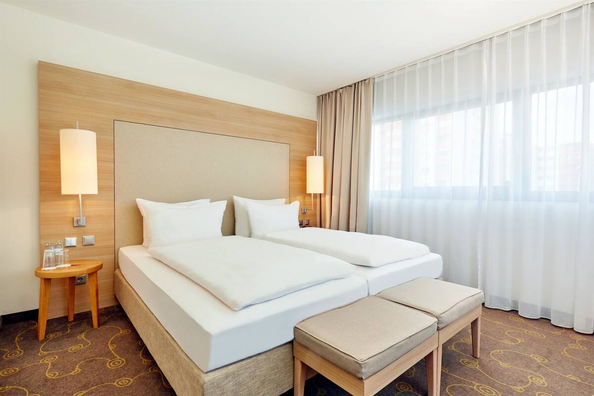 Habitación de hotel con dos camas, cabecero de madera, mesita de noche con lámpara, un banco y vista a la ciudad a través de cortinas traslúcidas.