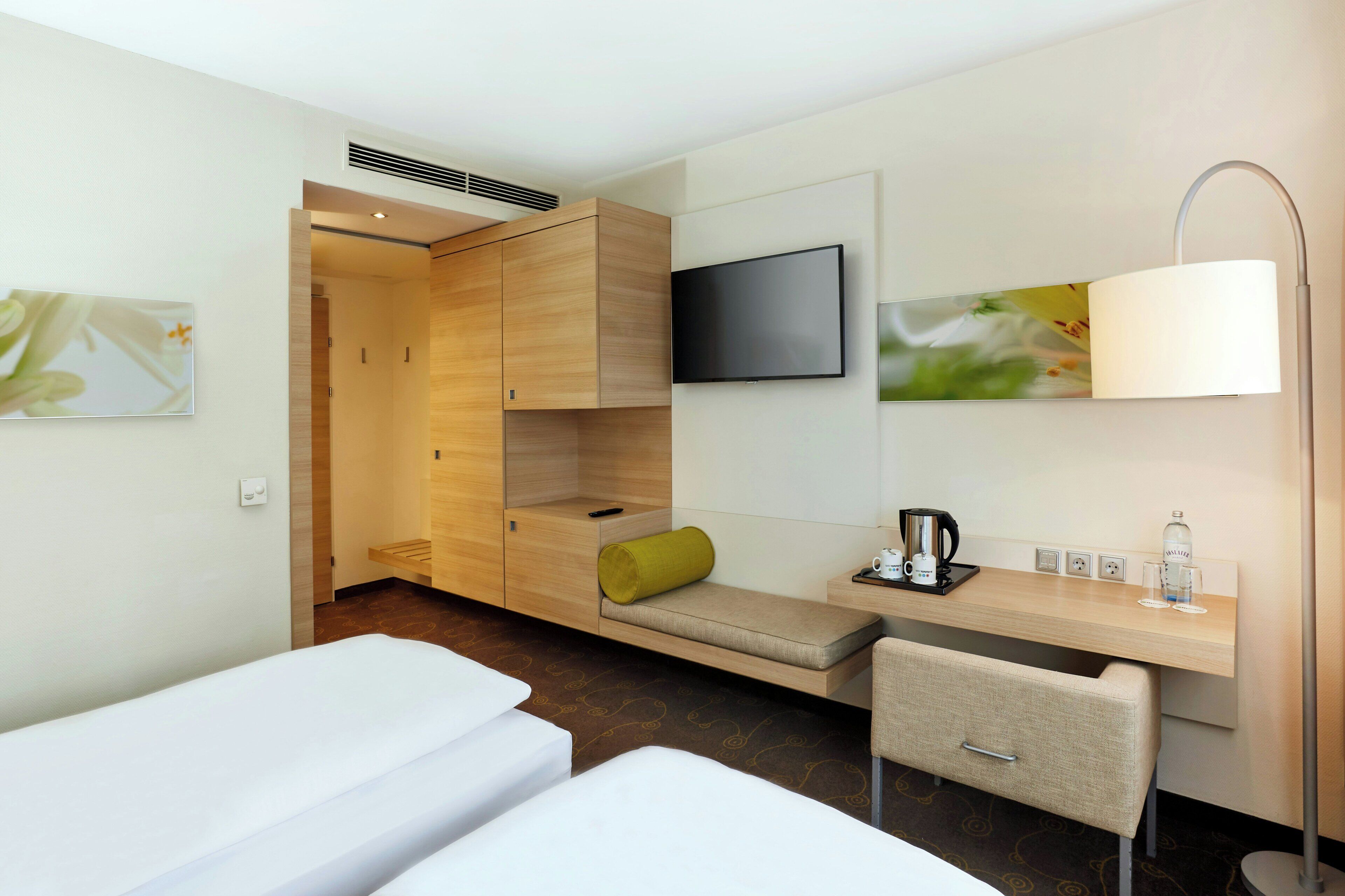 A hotel room with a bed, a desk, a chair, a TV, and a bedside table with a lamp and a bottle.