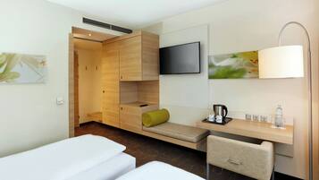 A hotel room with a bed, a desk, a chair, a TV, and a bedside table with a lamp and a bottle.