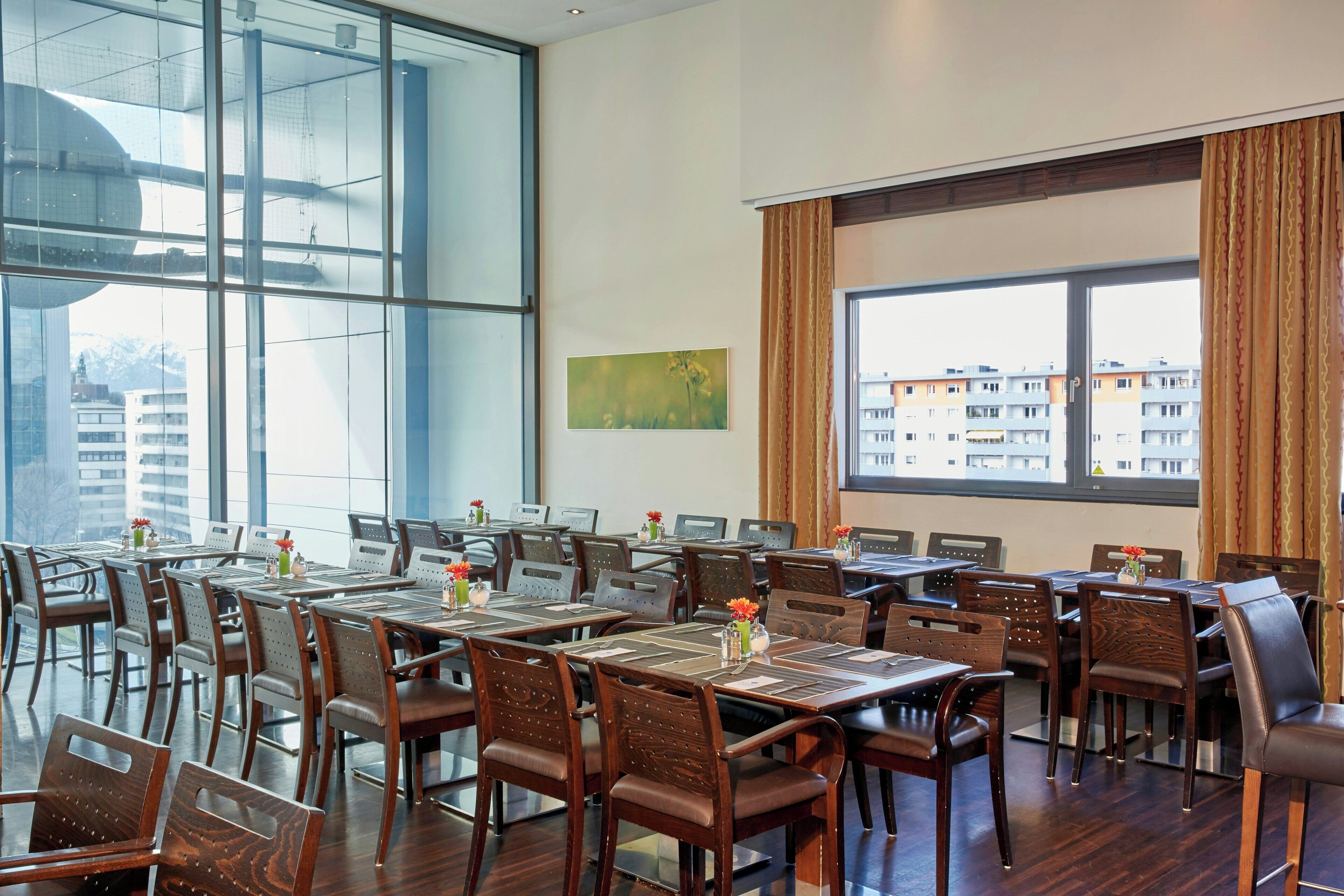 A conference room with multiple tables and chairs, large windows with curtains, and a painting on the wall.