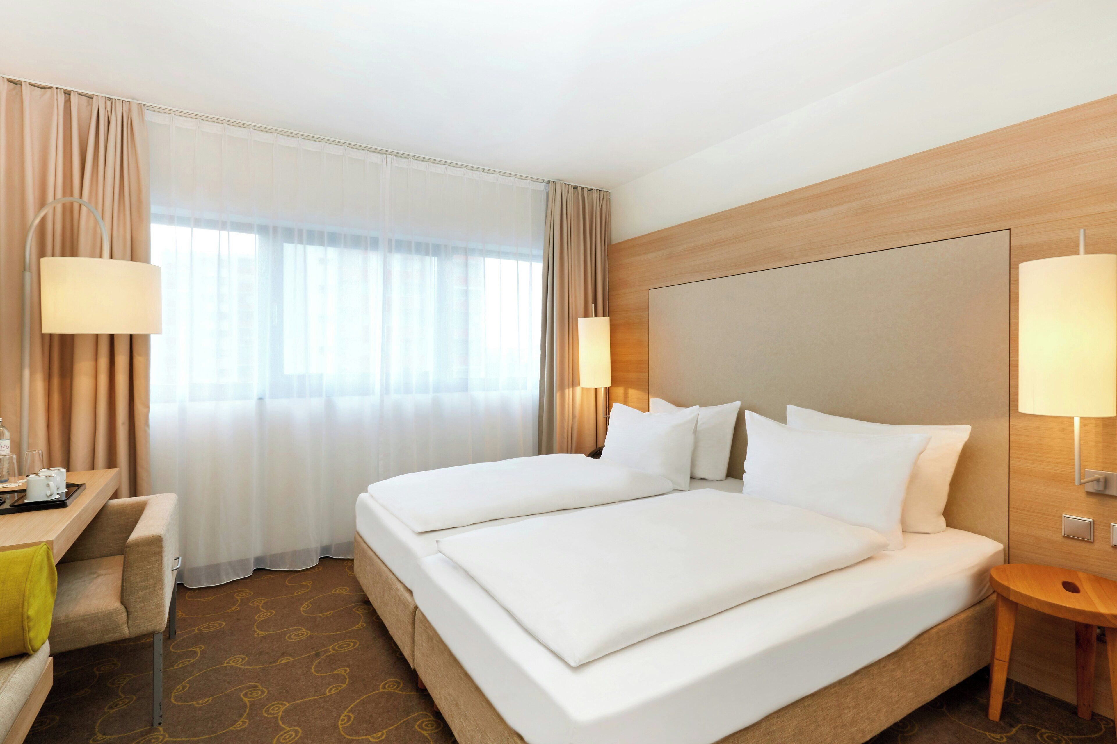 A hotel room with a large bed, two bedside tables, a desk with a chair, and a window with curtains.