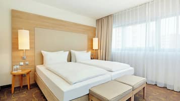 A hotel room with a double bed, a bedside table with a lamp, a bench, and a large window with curtains.