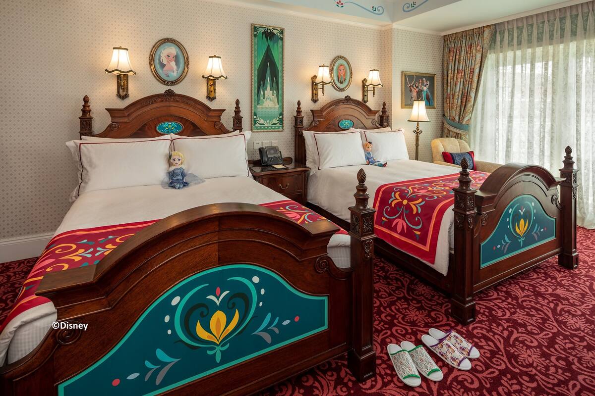 Ein Hotelzimmer mit zwei hölzernen Betten, jedes mit einem unverwechselbaren floralen Muster, einem Nachttisch mit Lampe und einem großen Fenster mit Vorhängen.