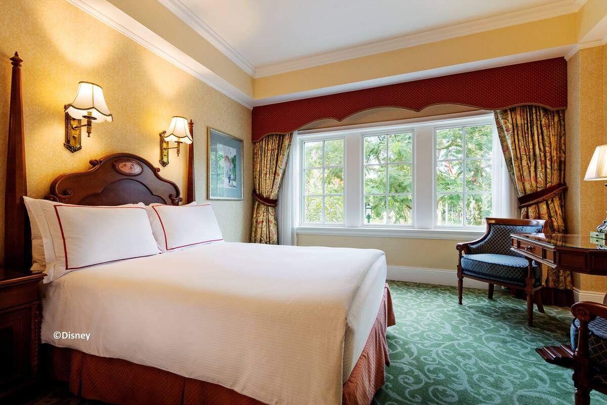 Ein Hotelzimmer mit einem großen Bett, einem Schreibtisch und einem Stuhl. Es gibt ein Fenster mit Vorhängen, eine Lampe und ein Bild an der Wand.