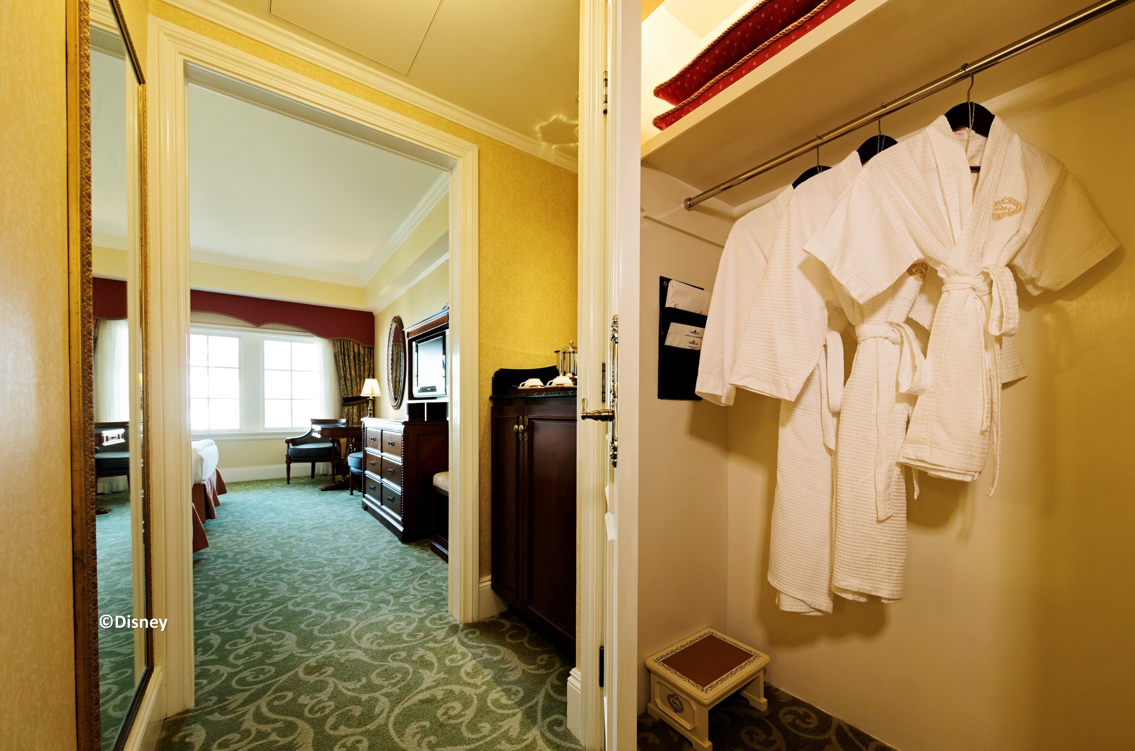 A hotel room with a green carpet, a bed, a dresser, and a mirror. There is a rack with white robes hanging on the wall.