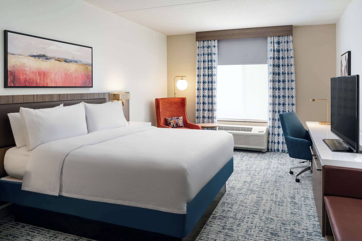 Ein Hotelzimmer mit einem großen Bett, einem Schreibtisch mit Fernseher, einem Sessel, einer Lampe und einem Fenster mit Vorhängen.