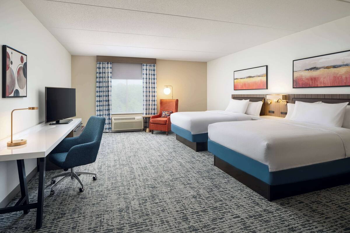 Ein Hotelzimmer mit zwei Betten, einem Schreibtisch, einem Sessel, einem Fernseher und Bildern an der Wand.