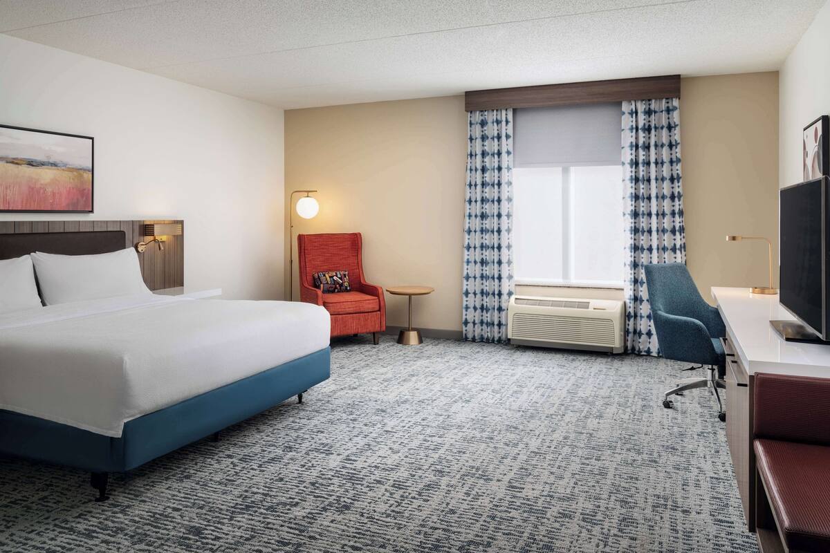 Ein Hotelzimmer mit einem großen Bett, einem Schreibtisch, einem Stuhl, einem kleinen Tisch und einem Fenster mit gemusterten Vorhängen.