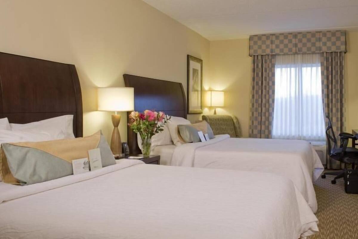 Ein Hotelzimmer mit zwei Betten, einem Schreibtisch, einem Stuhl, einer Lampe, einer Vase mit Blumen und einem Fenster mit Vorhängen.